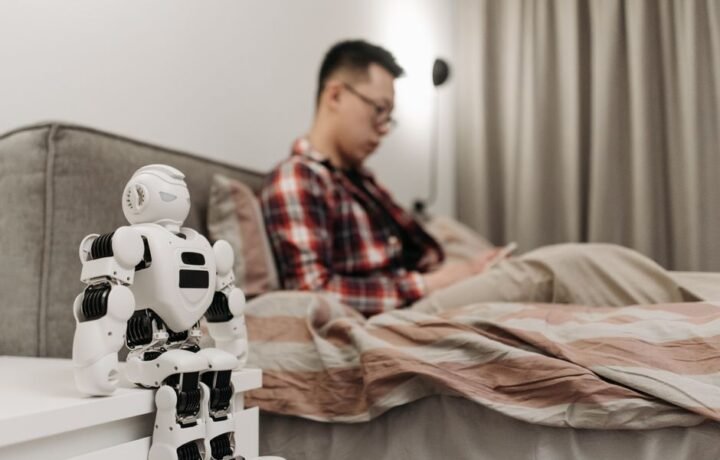 A modern bedroom featuring a robot on a bedside table and a man sitting on a bed.