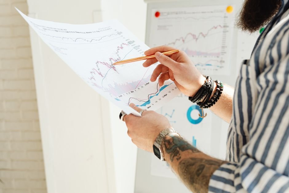 A man analyzing stock market charts with a pen, holding a paper report indoors.