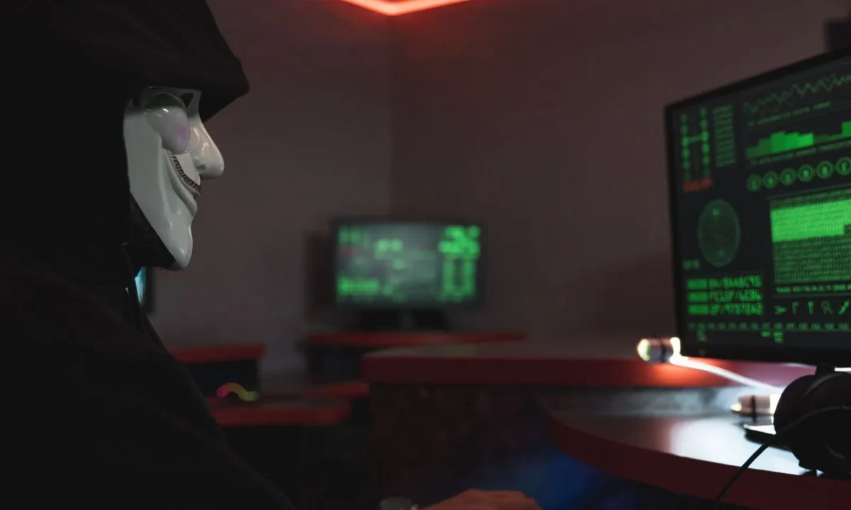 Hacker mascarado diante de um monitor em sala escura, representando a ameaça que a API anti-fraude ajuda a bloquear