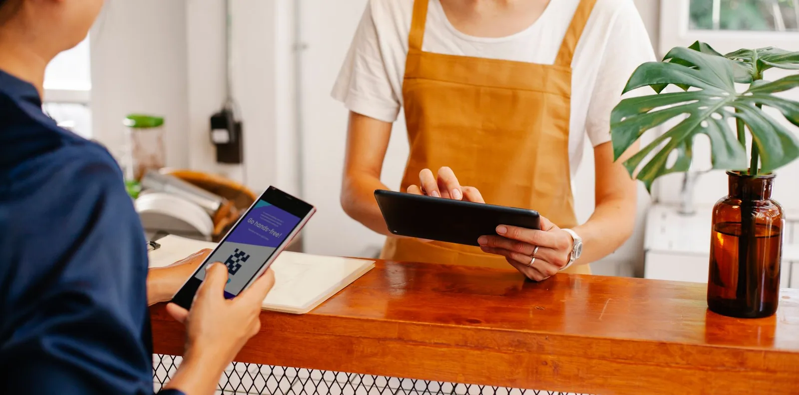 Atendente segurando um celular conversa com colega que usa um tablet no balcão de uma cafeteria