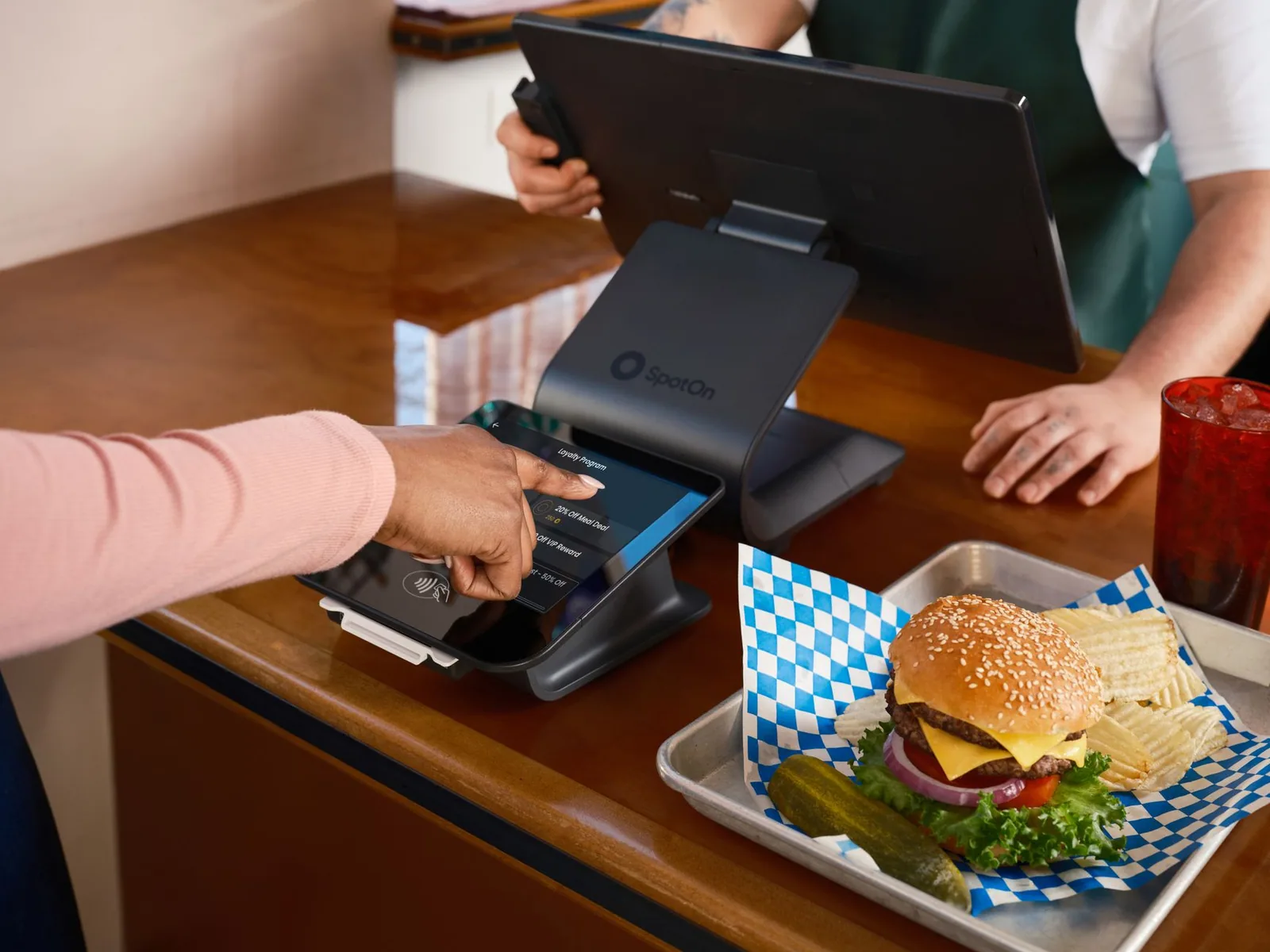 Balcão de restaurante com sistema POS SpotOn, hambúrguer servido e cliente interagindo no pagamento
